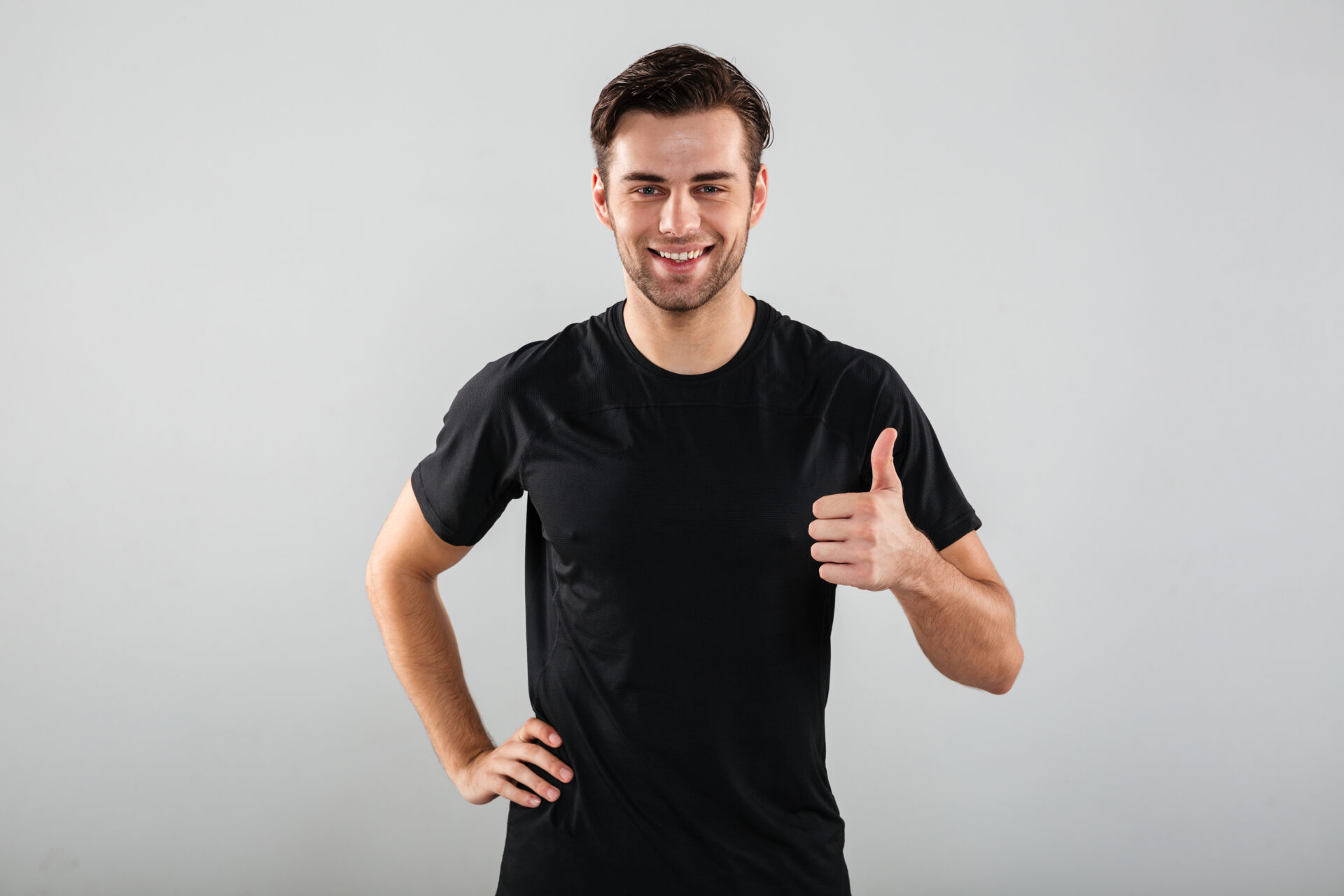 Photo of cheerful young sports man posing isolated over grey wall background. Looking camera showing thumbs up gesture.